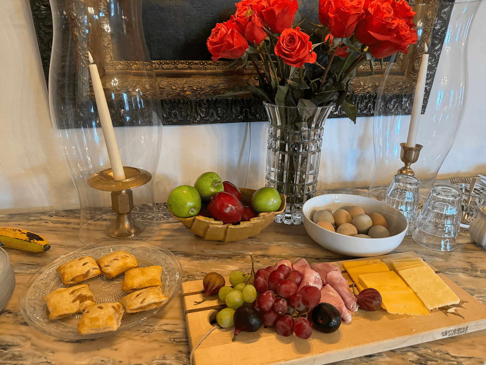 A table arranged with pastries, assorted fruits, cheeses, meats, and a vase of red roses.