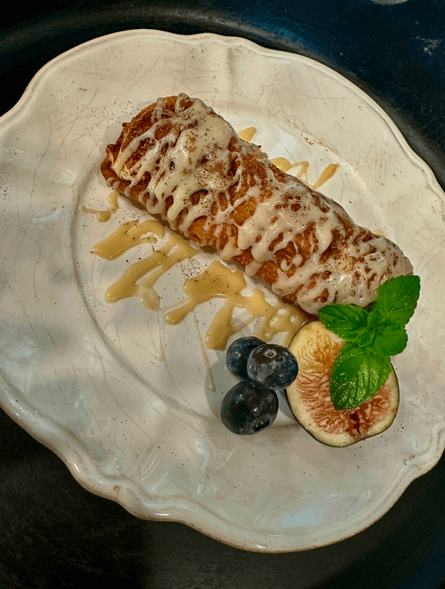 A dessert featuring a cream-drizzled pastry on a plate, accompanied by blueberries and a fig slice, garnished with mint.