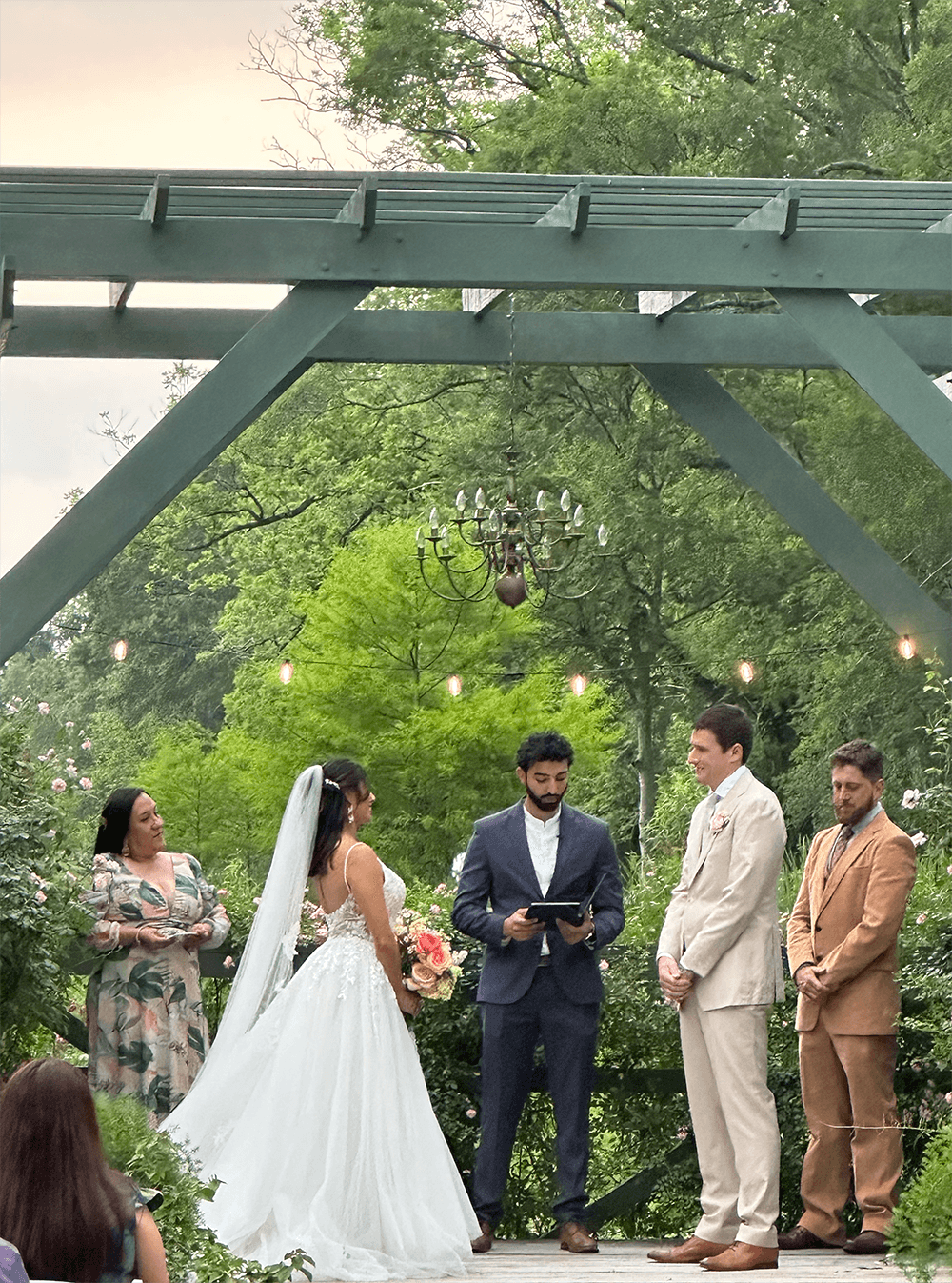 A wedding ceremony takes place outdoors under a green arbor with four attendees, including the bride in a white gown and a man officiating.