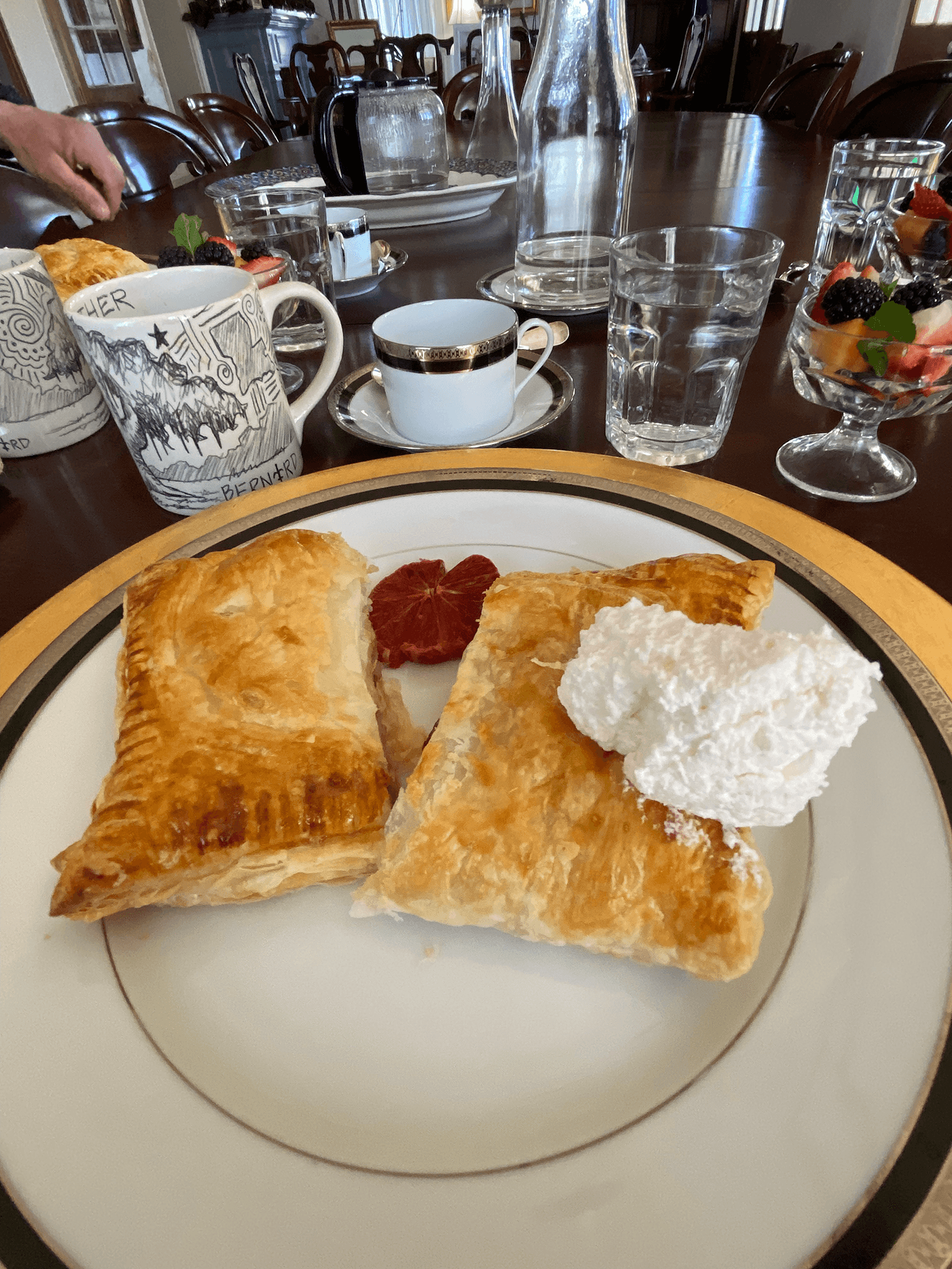 A plate with two flaky pastries, a slice of citrus, and a dollop of whipped cream, accompanied by various drinks and fruit in the background.
