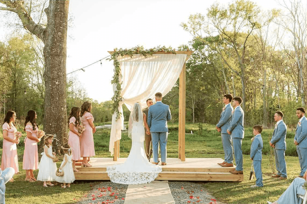 A wedding ceremony taking place outdoors under a floral-draped arch with guests gathered around.