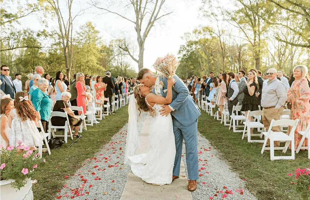 Bride and groom share a kiss on a flower-strewn aisle, surrounded by smiling guests seated in rows outdoors, creating a joyful and celebratory scene.